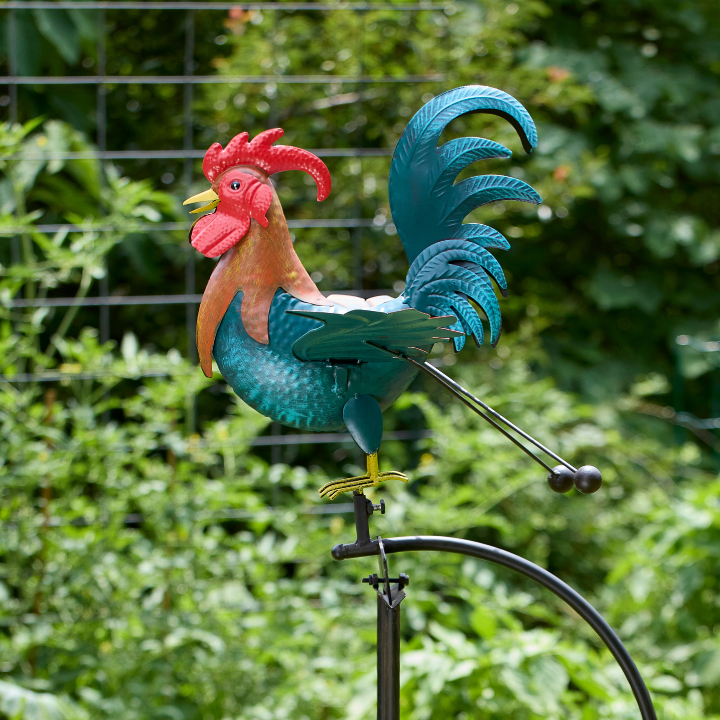 Close-up of hand-painted teal metal rooster with bold red comb and tail feathers, perched on balance rocker mechanism against blurred green garden background