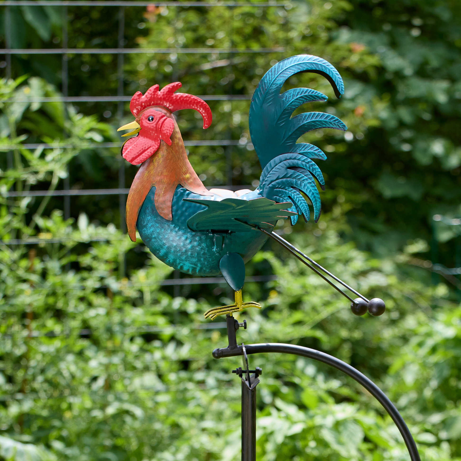 Close-up of hand-painted teal metal rooster with bold red comb and tail feathers, perched on balance rocker mechanism against blurred green garden background