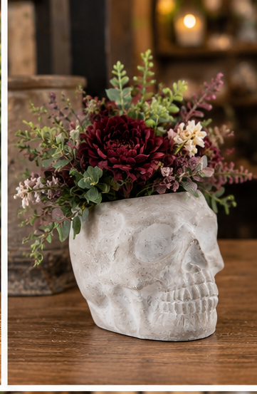 Side view of a gray cement sugar skull planter styled with burgundy flowers and greenery on a wooden surface