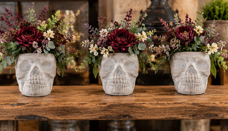 Three gray cement sugar skull planters styled with burgundy and cream flowers on a rustic wooden shelf