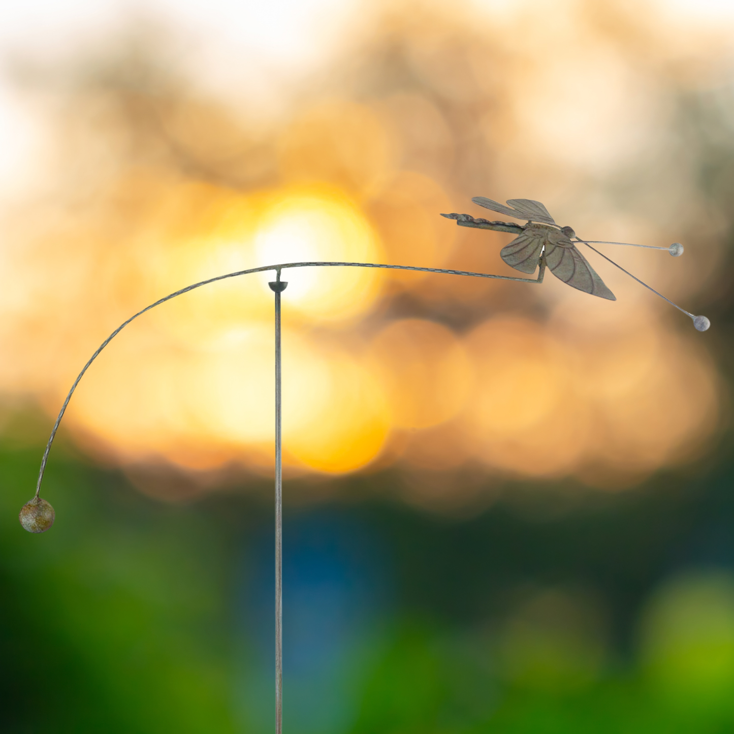 Kinetic dragonfly wind art balancer garden stake silhouetted against warm golden sunset bokeh background
