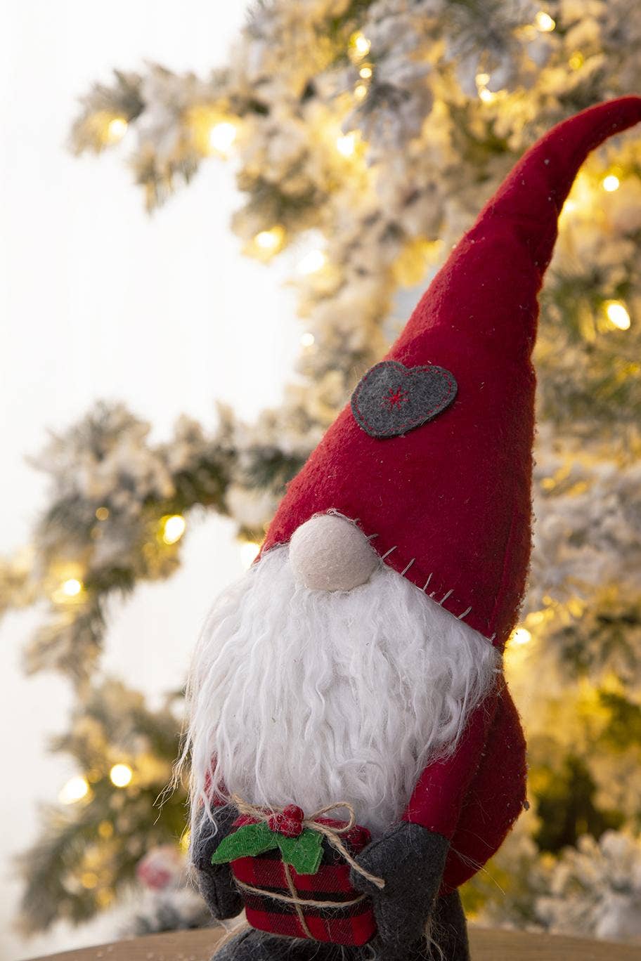 Close-up of the Christmas gnome’s curved red hat with gray heart patch, fluffy white beard, and red gift with twine and holly accents.