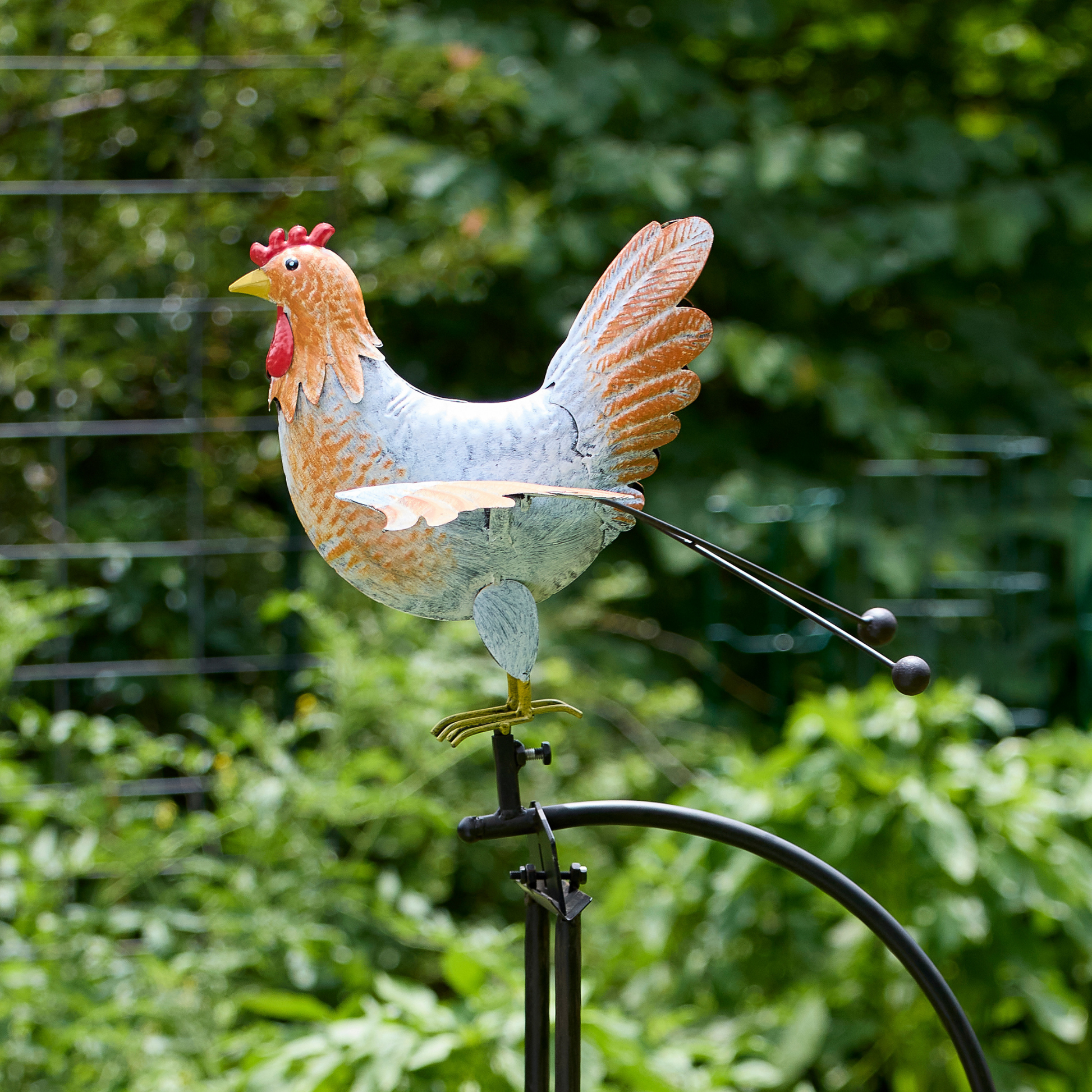 Close-up of hand-painted metal hen balance rocker with wings spread and red comb detail against blurred green garden background