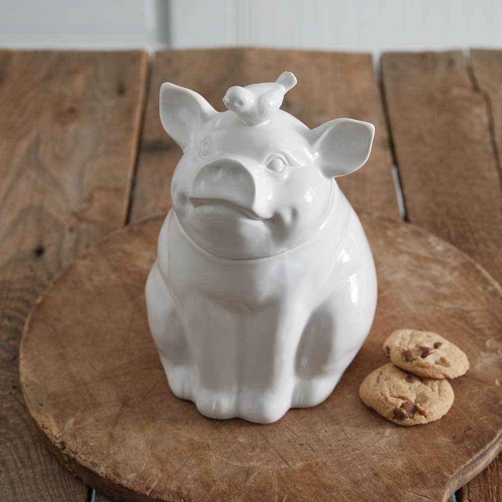 White ceramic piglet cookie jar with its lid on, displayed on a wooden board beside two cookies.
