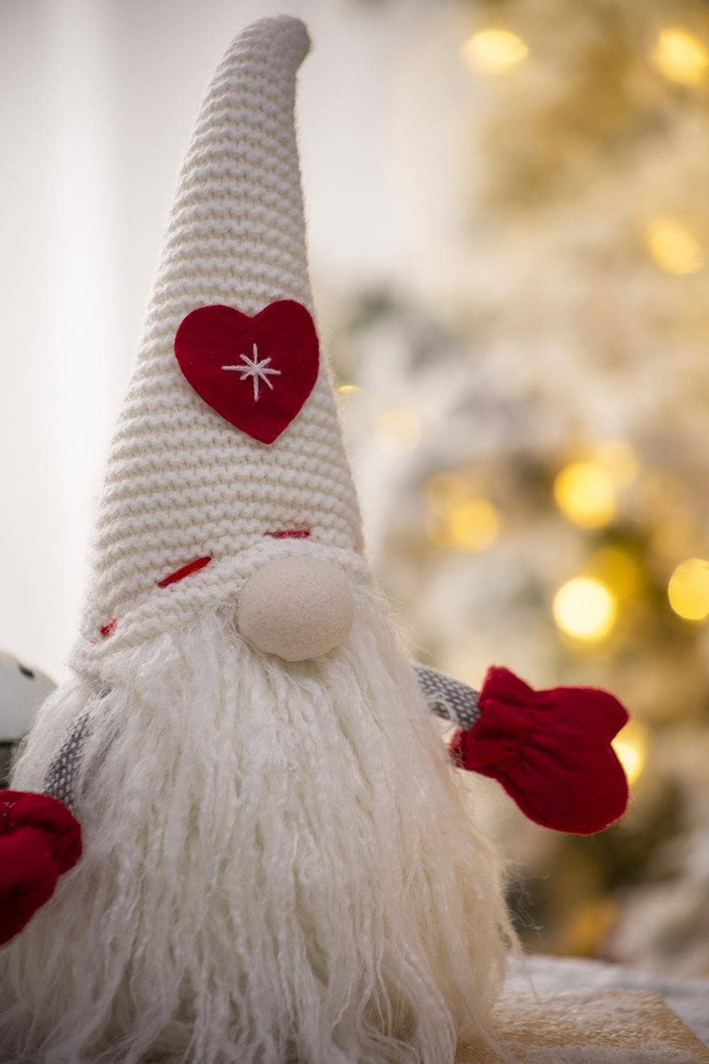 Close-up of the Christmas gnome’s white knit hat with a red heart and stitched white starburst, cream nose, fluffy white beard, and red mittens.