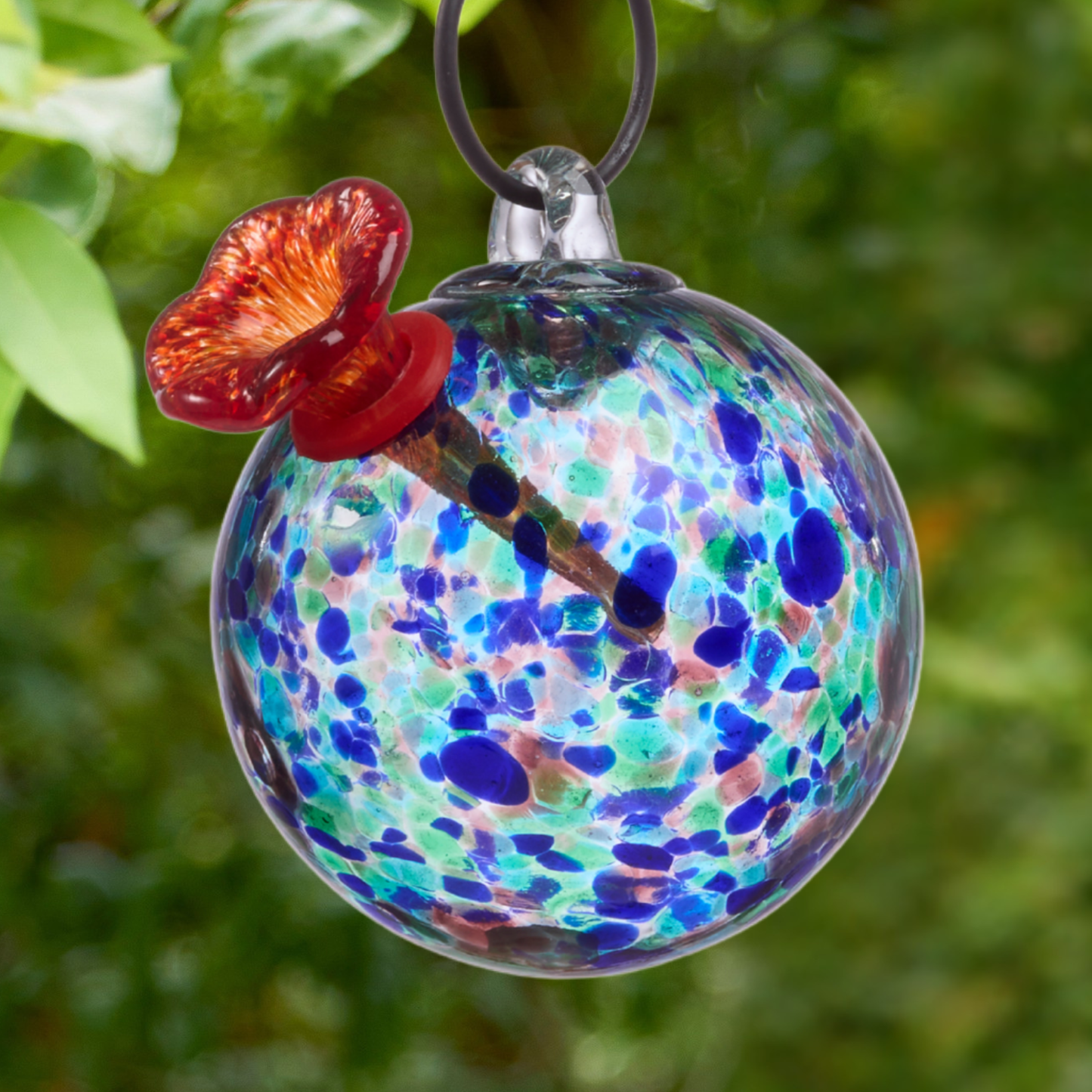 Close-up of hand-blown turquoise art glass hummingbird feeder globe with blue and teal speckle pattern and red glass flower feeding port against green foliage