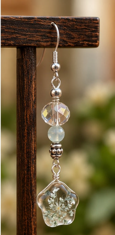 Side view of a floral dangle earring with clear faceted beads and a white flower design hanging from a wooden stand