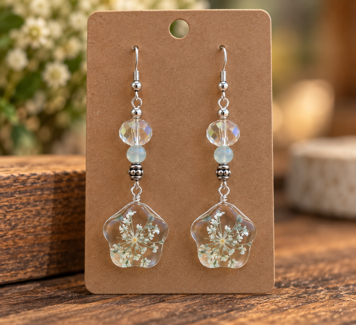 Handmade white floral dangle earrings with clear flower-shaped pendants and pale blue bead accents displayed on a kraft card
