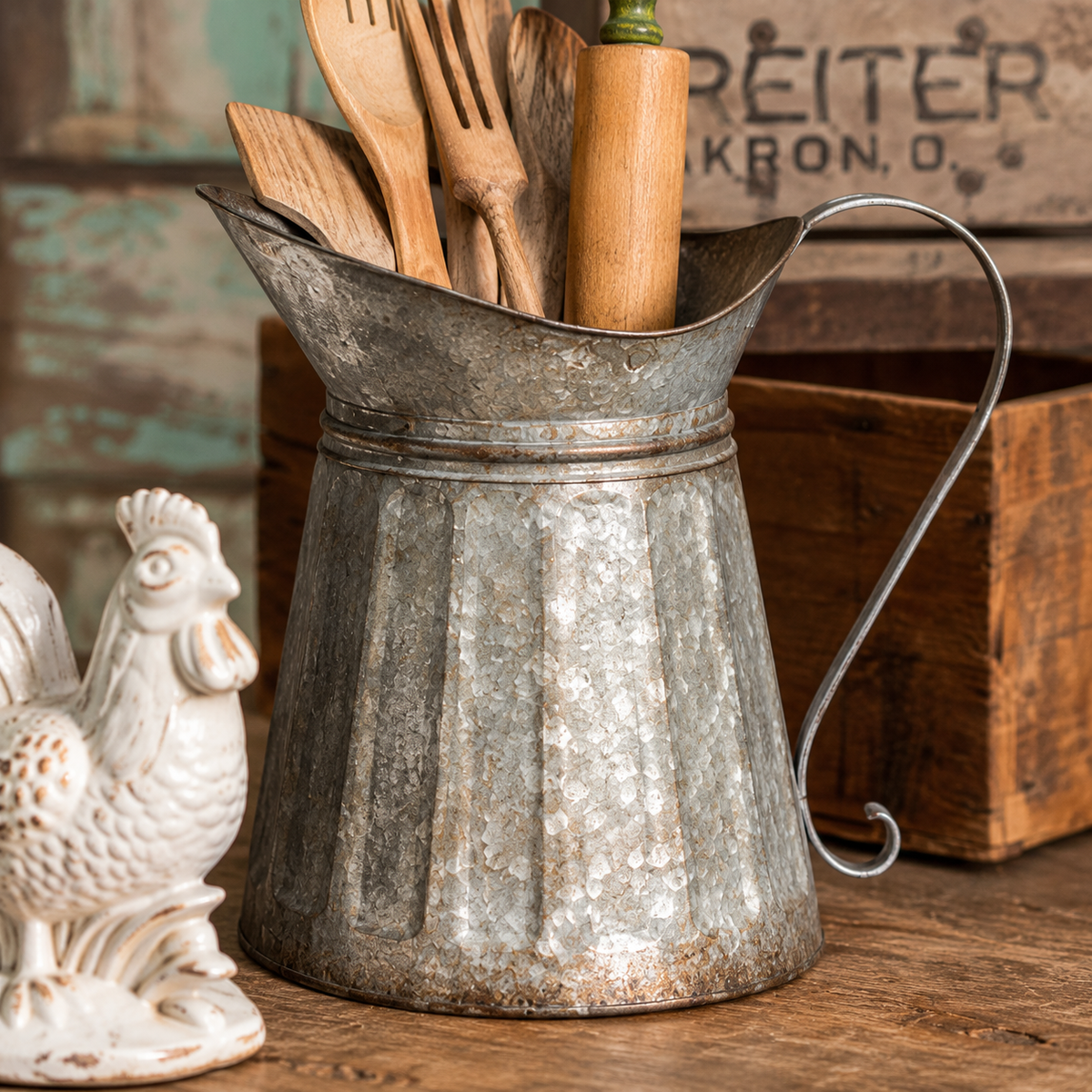Galvanized metal milk pitcher holding wooden kitchen utensils beside a ceramic rooster on a rustic wooden surface