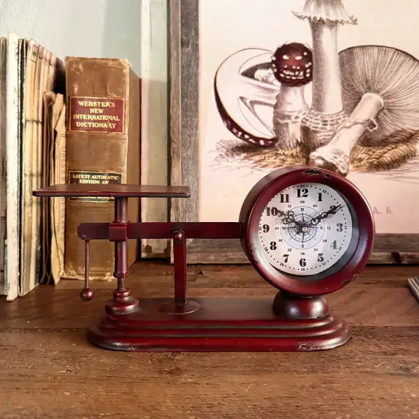 Red balance scale tabletop clock displayed in front of vintage books and framed mushroom artwork