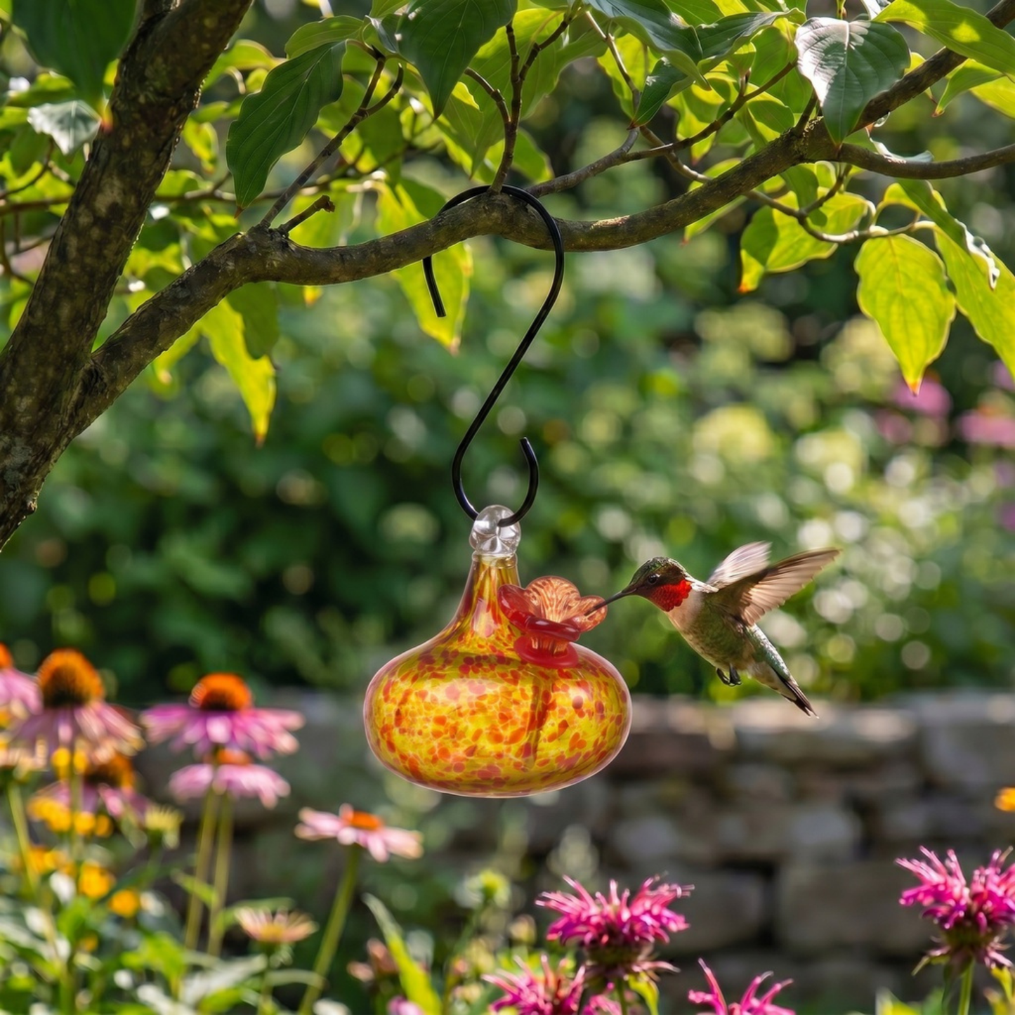 Hand-blown yellow art glass hummingbird feeder hanging outdoors in a garden
