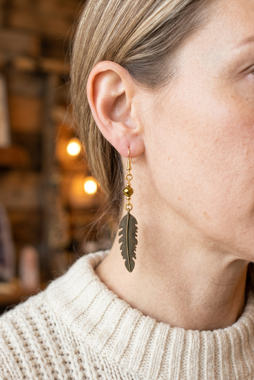 Woman wearing a feather dangle earring with a dark antiqued feather charm, amber-gold faceted bead, and gold-plated hook